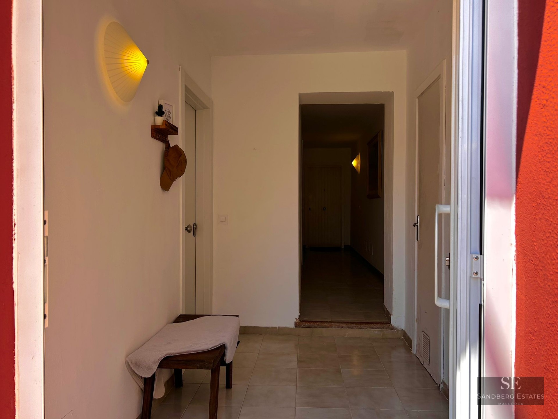 A clean white hallway with a wooden bench, tiled floor, and a glowing fan-shaped wall light near multiple white doors.