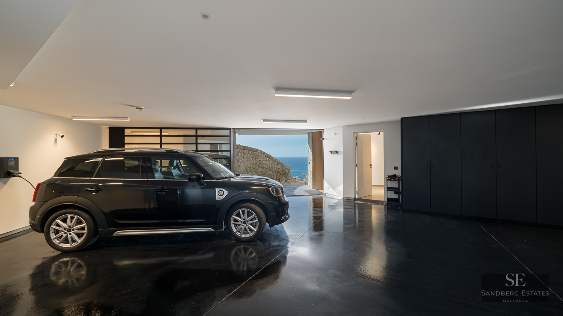 A black car parked in a modern garage with a glossy black floor, EV charger, and a sea view in the background.