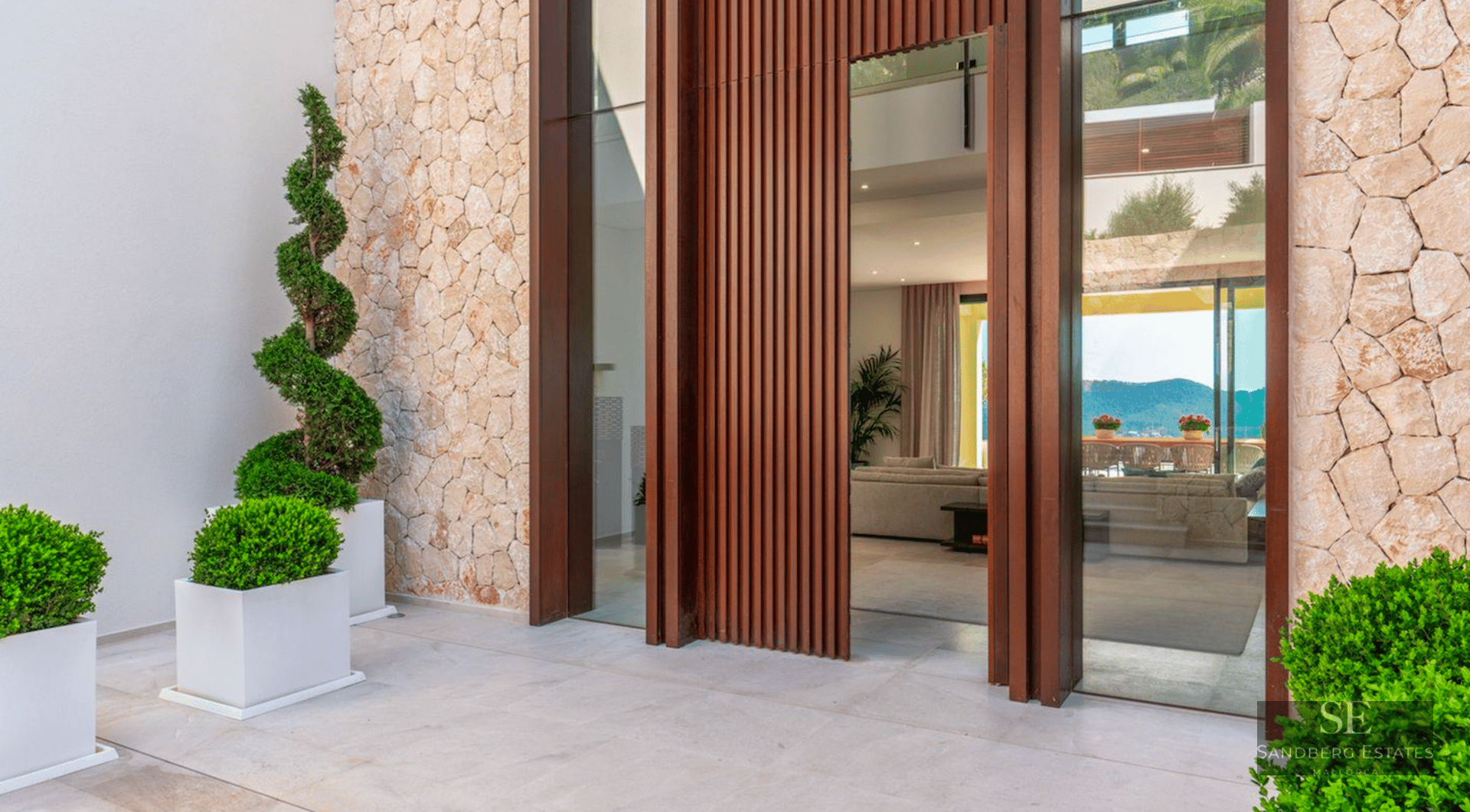 Grand villa entrance featuring a massive slatted wood door, natural stone walls, and spiral topiary in white planters.