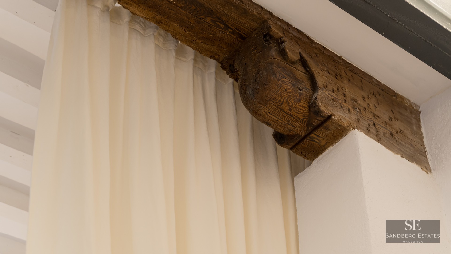 Close-up of a hand-carved wooden beam bracket and soft white curtains against a white plastered wall.