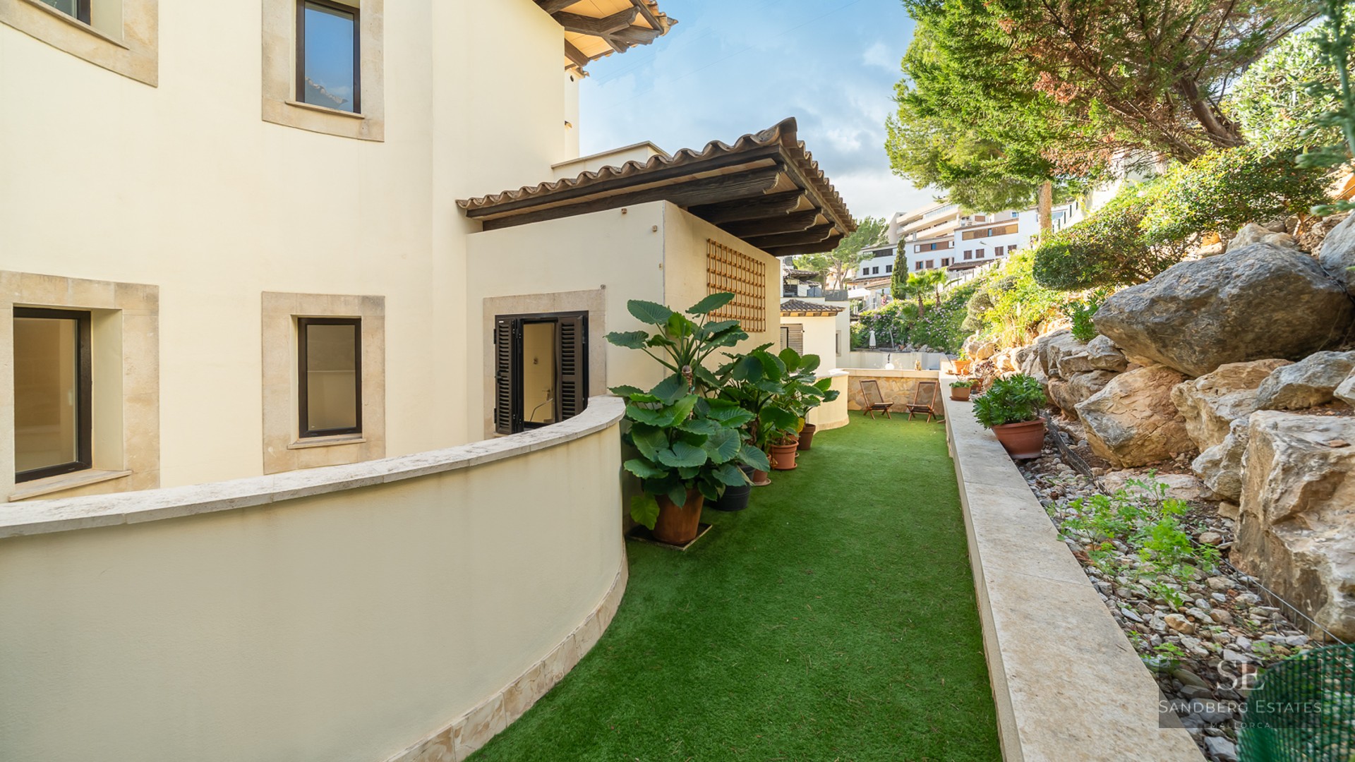 Curved villa wall with artificial grass path, potted tropical plants, and a natural stone rockery garden.