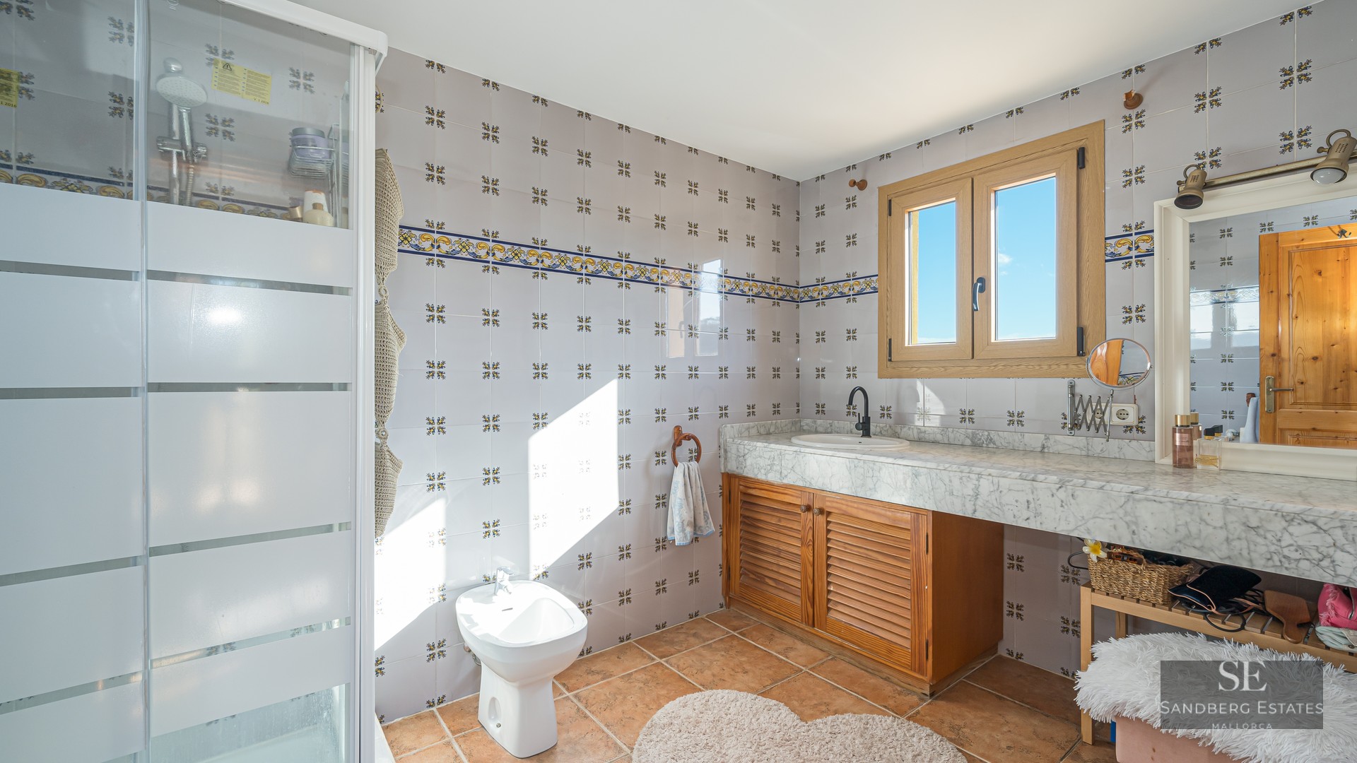 Bright bathroom featuring decorative wall tiles, white marble countertop, wooden vanity, bidet, and terracotta flooring.
