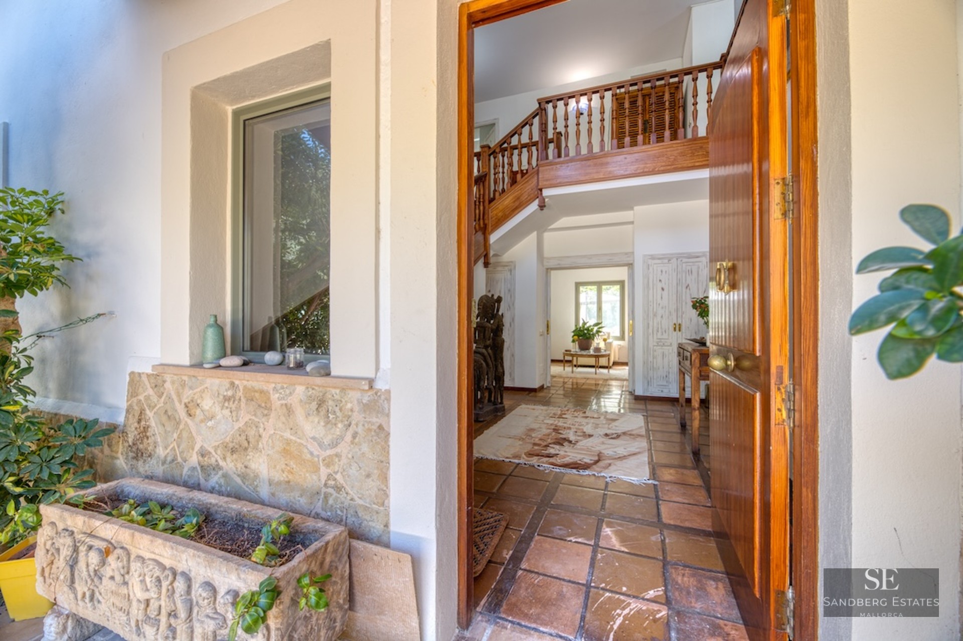 Open wooden door looking into a Mediterranean hallway with terracotta tiles and a wooden staircase.