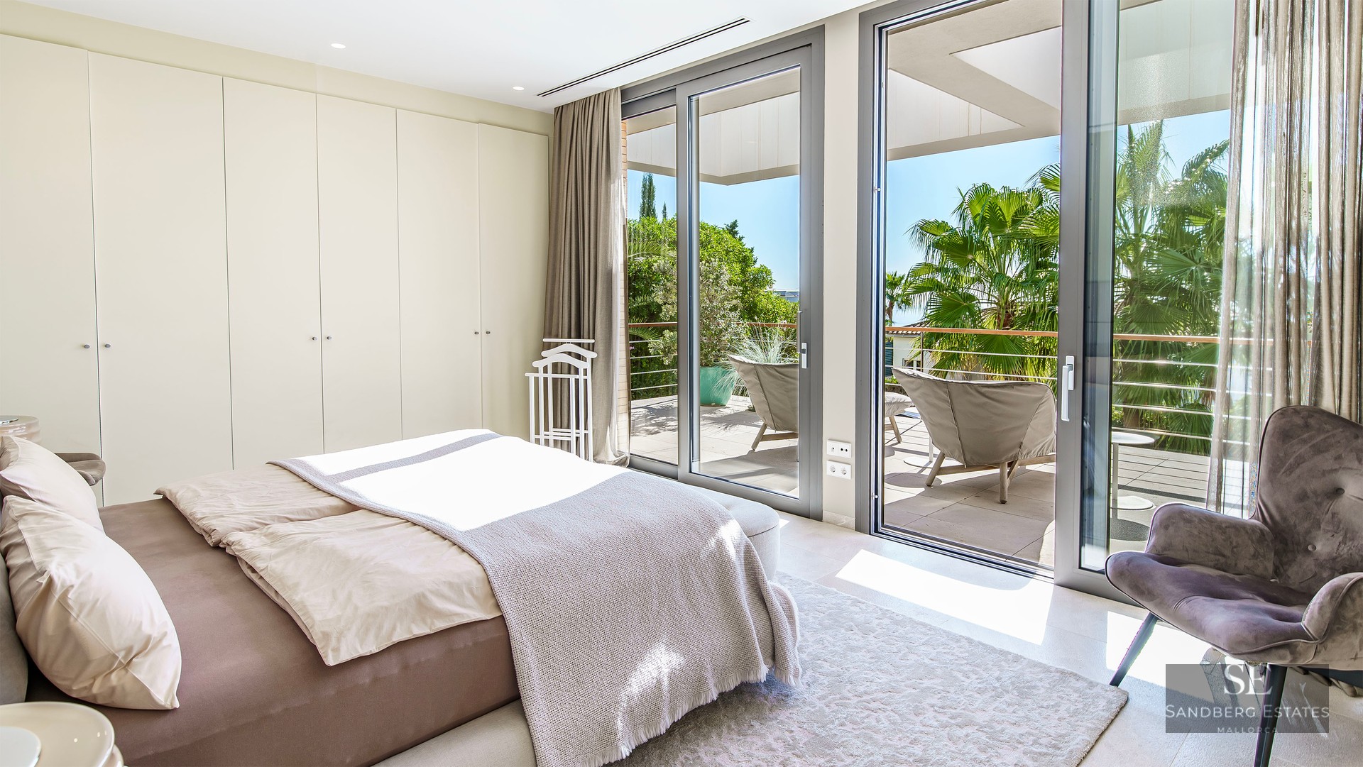 Chambre moderne avec baies vitrées donnant sur une terrasse ensoleillée et des palmiers.
