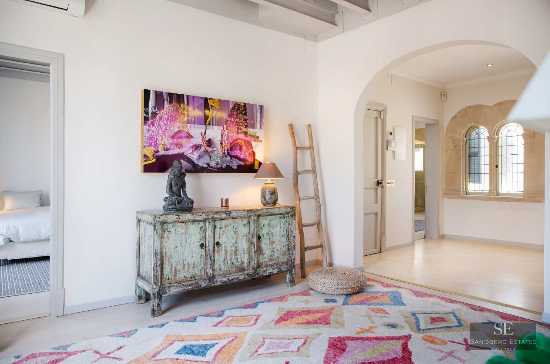 Interior hallway featuring a colorful geometric rug, vintage turquoise sideboard, rustic ladder, and arched stone windows.
