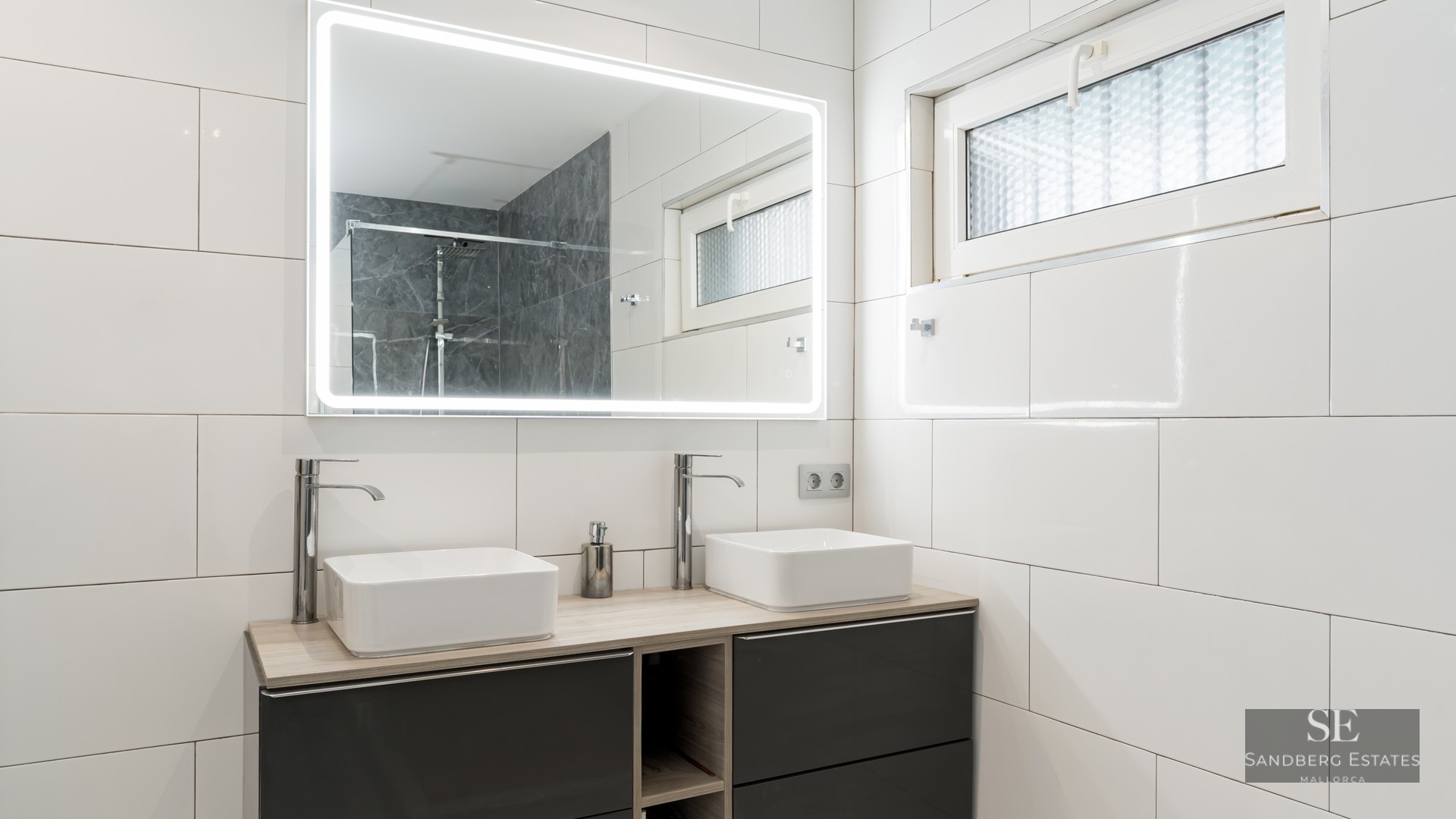Salle de bain moderne avec deux vasques carrées, un grand miroir éclairé et des murs en carrelage blanc.