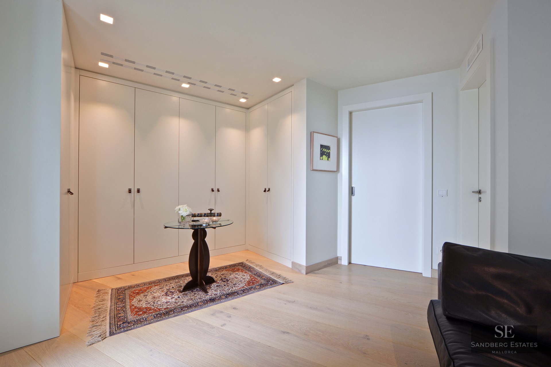 Bright entrance hall with white built-in closets, light wood floors, a decorative rug, and a glass side table.