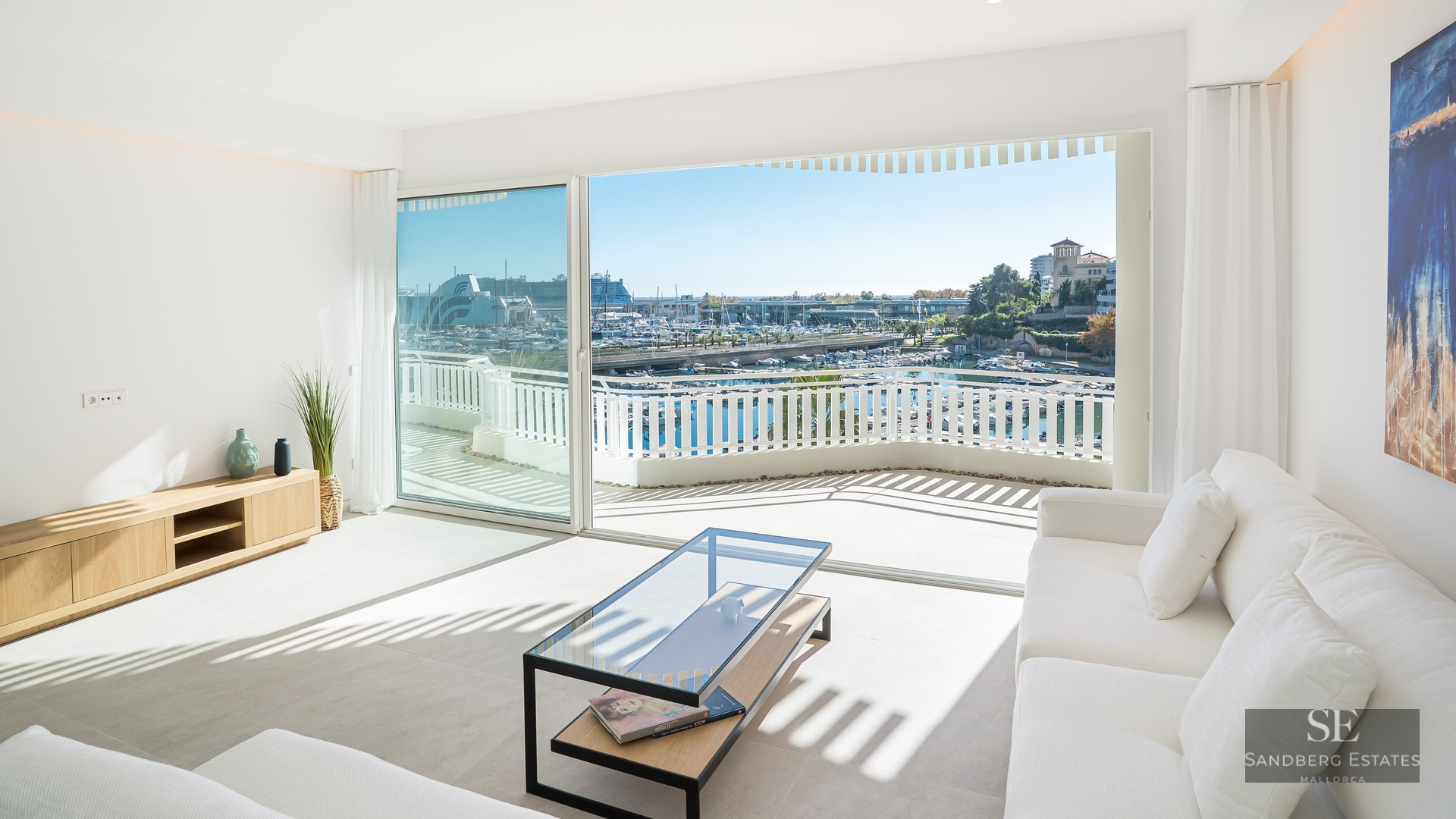 Bright minimalist living room with white sofas and large glass doors opening to a harbor view and terrace.