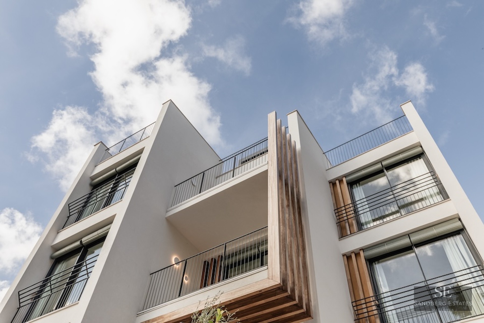 Low-angle view of a modern white building facade with wooden accents and black balcony railings against a blue sky.