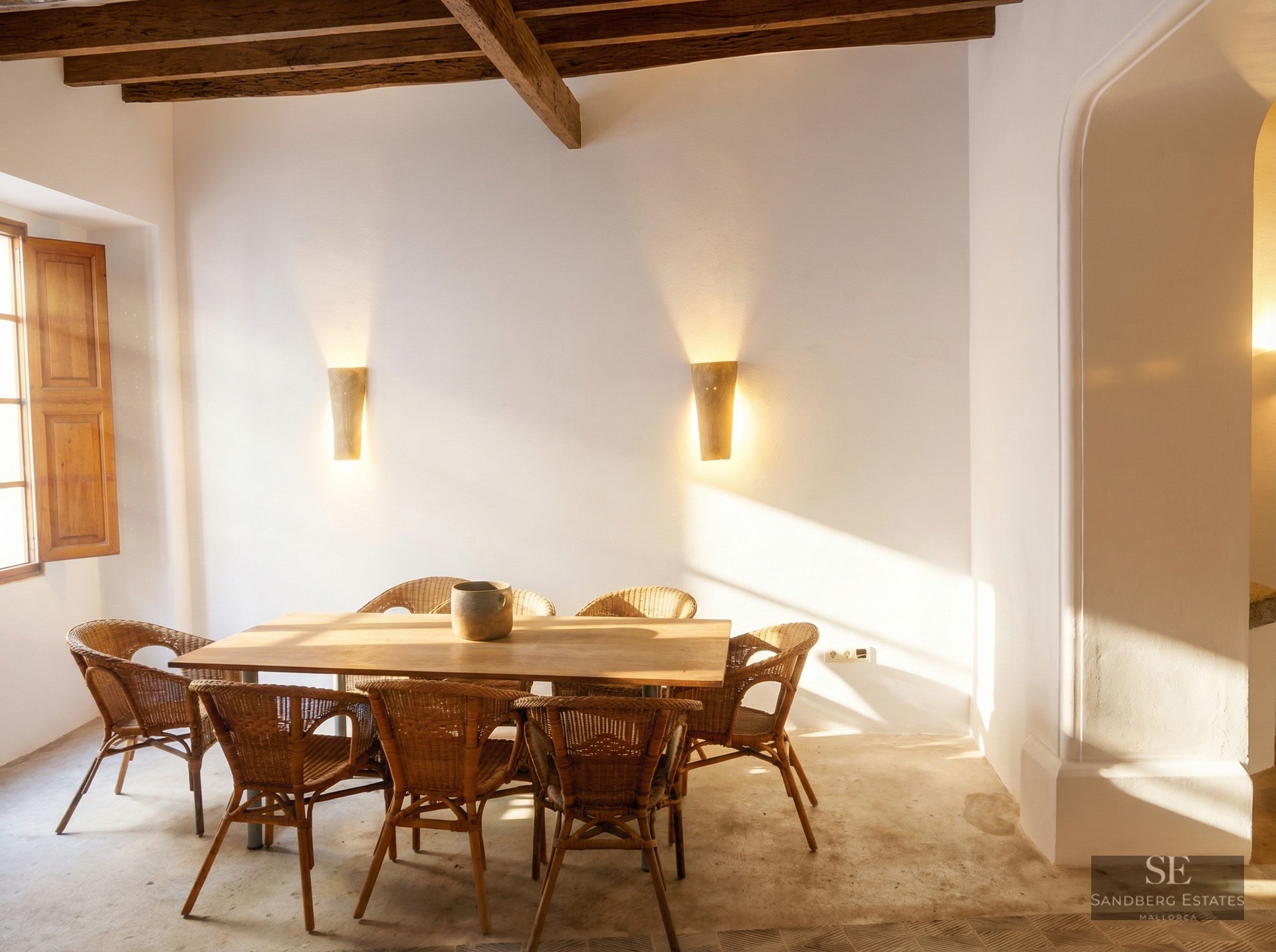 A wooden dining table with wicker chairs in a white room featuring dark ceiling beams and warm sunlight.