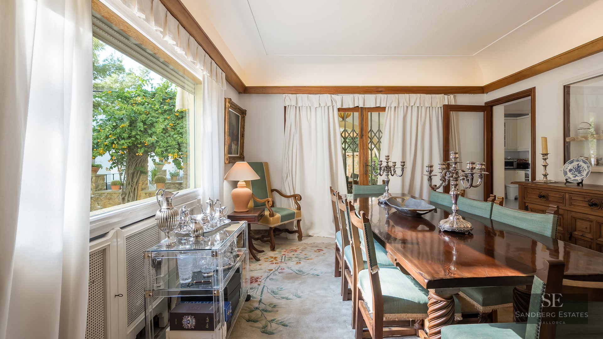 Formal dining room featuring a polished wood table, green upholstered chairs, and silver candelabras by a garden window.