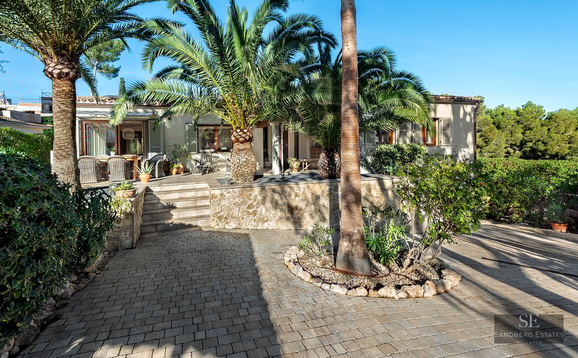 Exterior of a Mediterranean house with stone walls and large palm trees under a clear blue sky.
