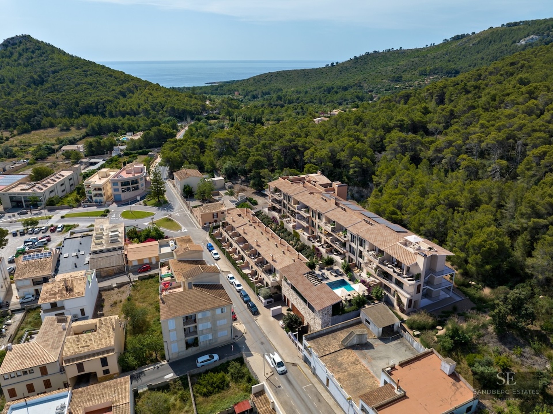 Drone shot of Mediterranean apartment buildings with a swimming pool, surrounded by green forest and sea horizon.