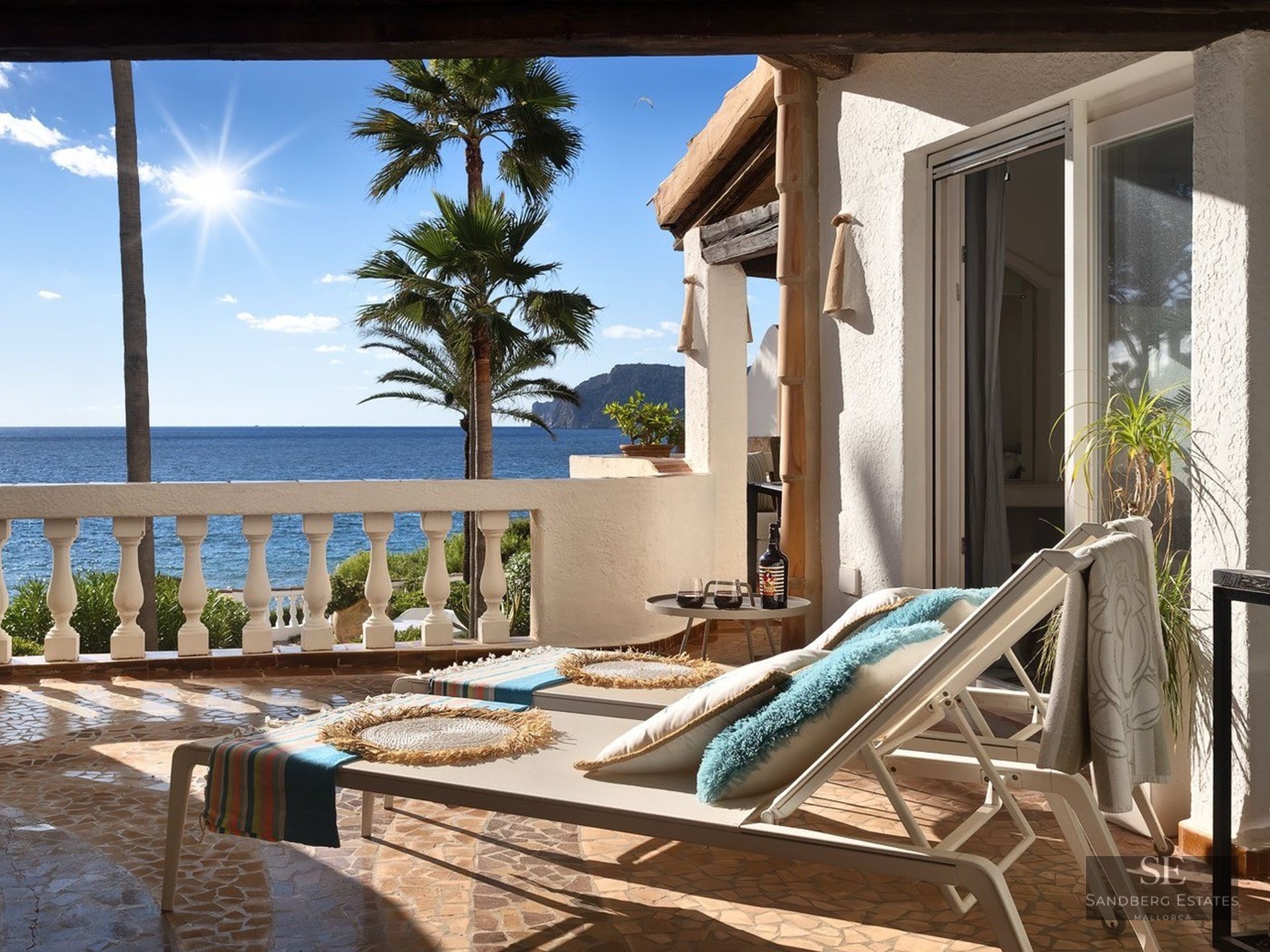 White sun loungers on a Mediterranean terrace overlooking the ocean with palm trees under a bright sun.