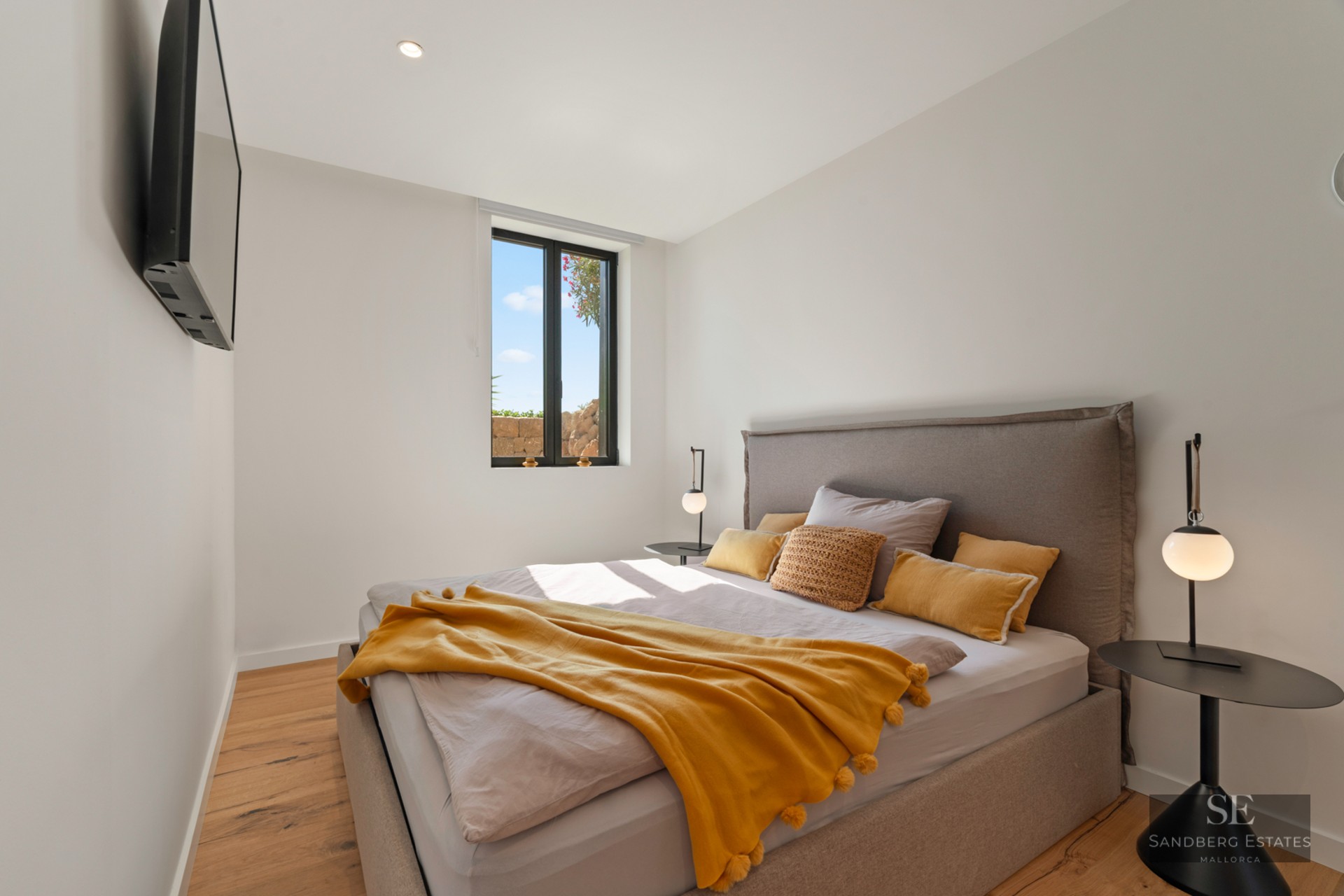 Modern bedroom with a grey bed, mustard yellow throw, wooden floors, and a window showing a bright sky.