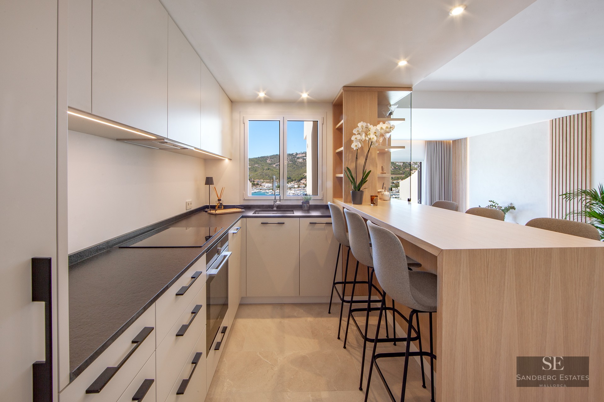 Modern kitchen with white cabinets, dark countertops, a wooden breakfast bar with three stools, and a window view of a harbor.