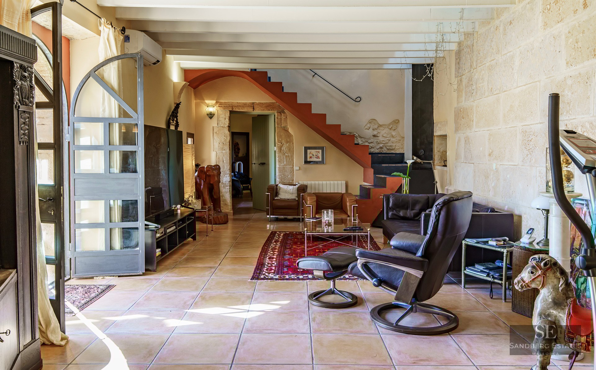 Sun-drenched living room with natural stone walls, terracotta tiles, exposed beams, and leather lounge chairs.