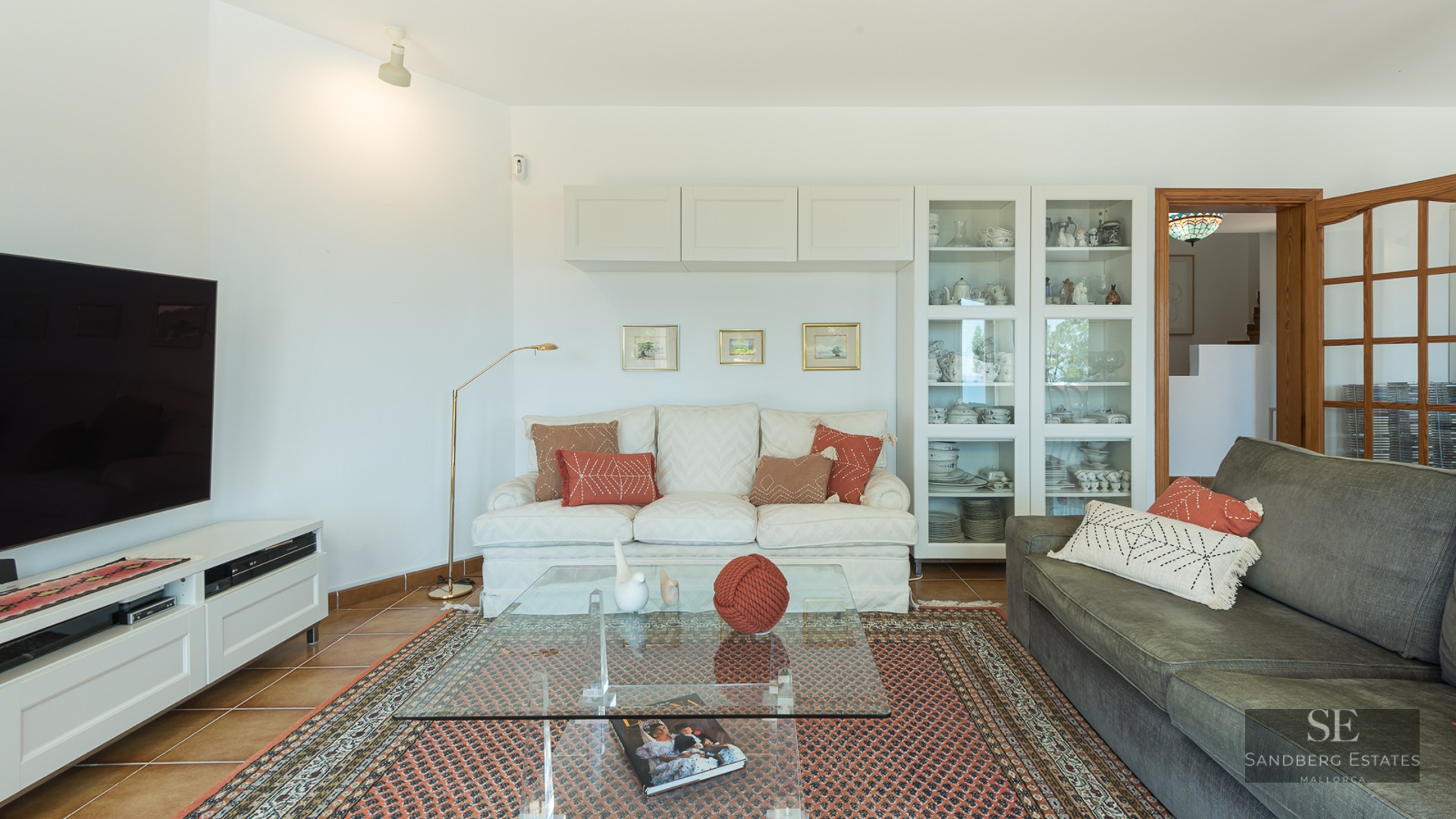Bright living room with sofas, glass coffee table on an ornate rug, large TV, and terracotta tile floors.
