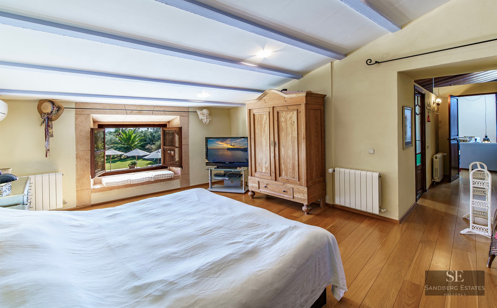 A cozy bedroom with wooden floors, a large bed, rustic armoire, and a window overlooking a lush garden.