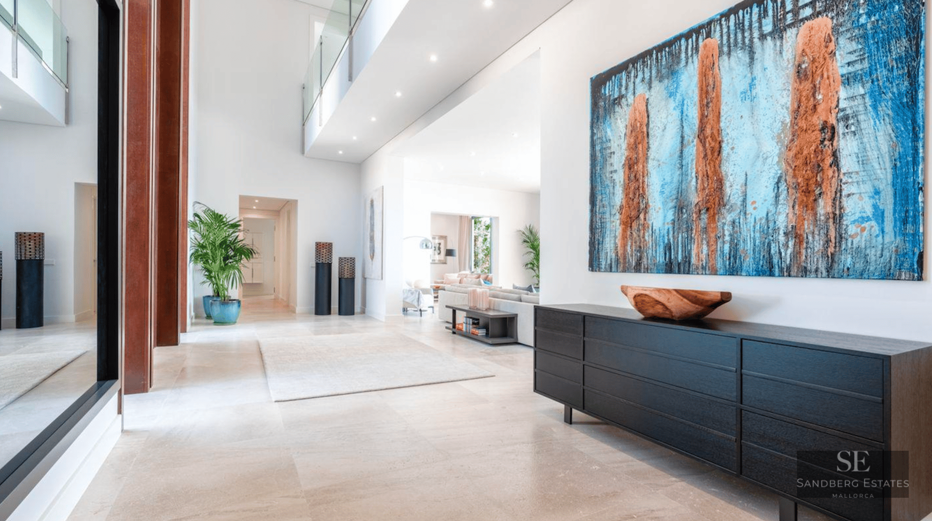 Bright entrance hall with stone floors, a black sideboard, a large blue abstract painting, and a glass mezzanine.