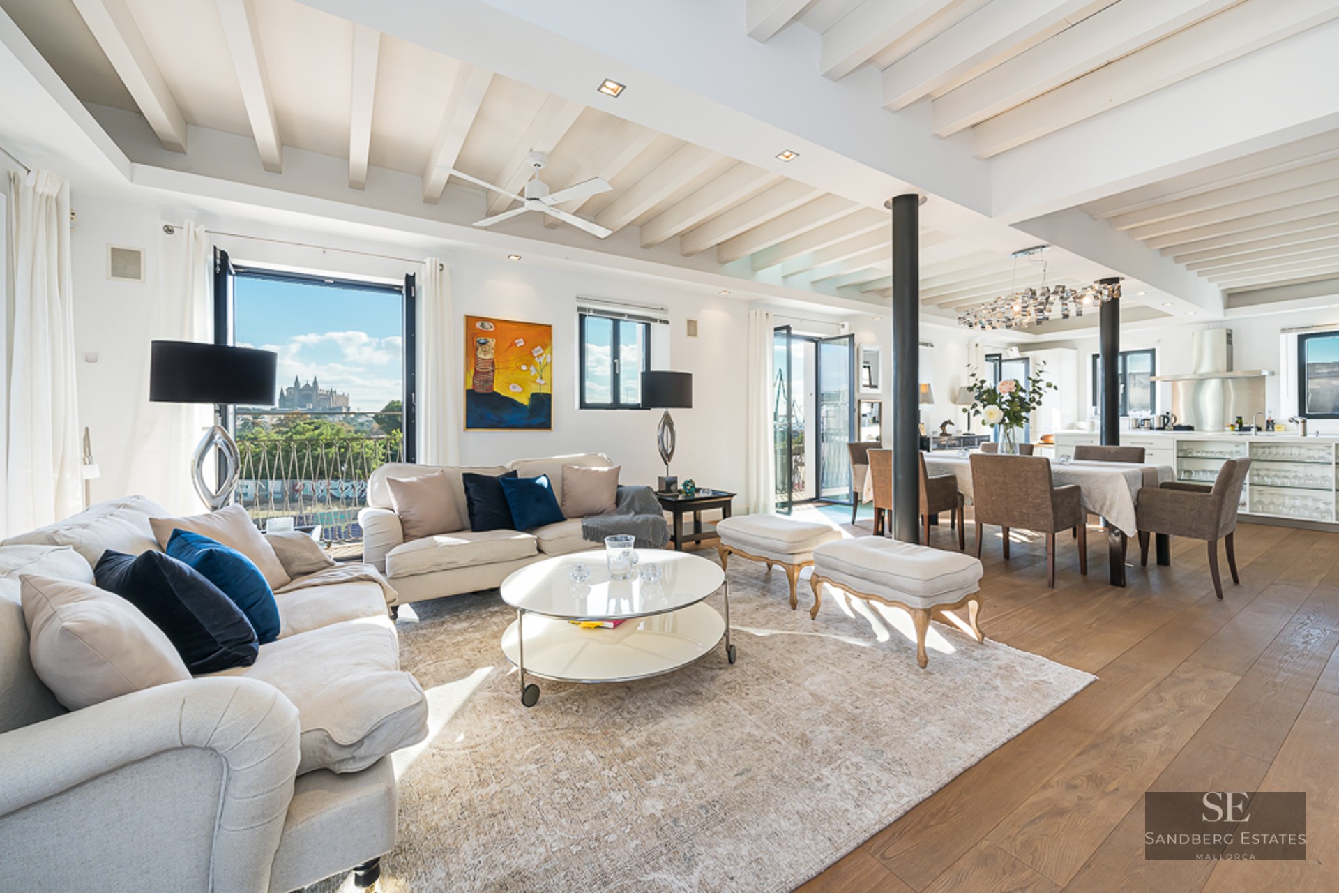 Spacious white living room with beamed ceilings, wooden floors, and a view of a cathedral through large windows.