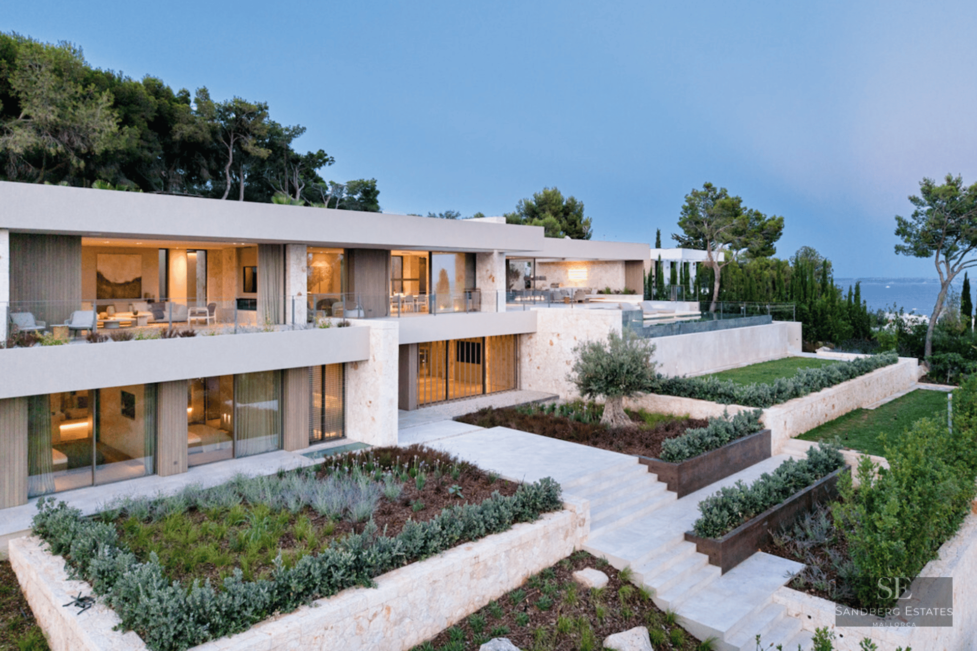 Modern multi-level villa with stone walls, large windows, and tiered Mediterranean gardens overlooking the sea at dusk.