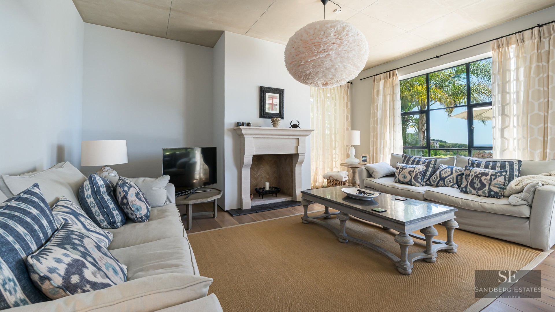 Spacious living room with beige sofas, stone fireplace, and large windows viewing garden palms.