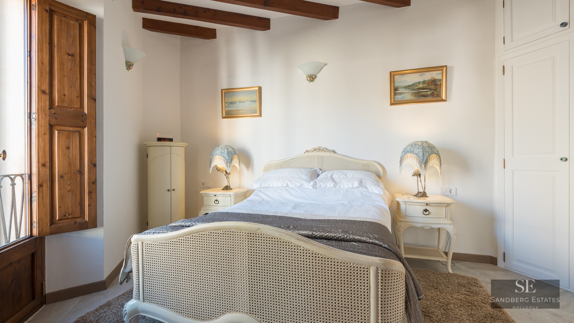 Cozy bedroom featuring a rattan bed, wooden ceiling beams, bedside lamps, and dark wood window shutters.