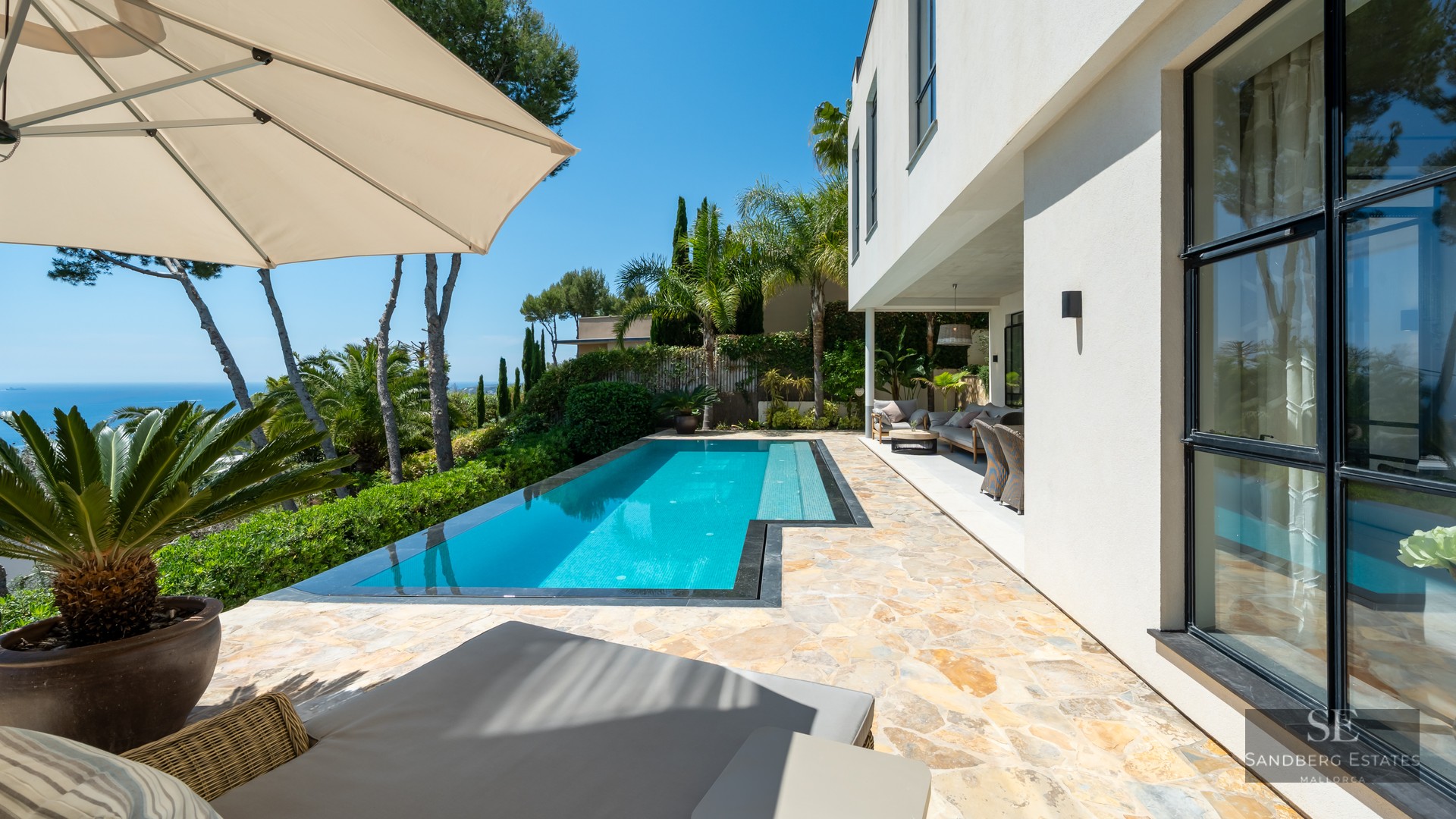 A turquoise swimming pool on a stone patio next to a white modern villa, overlooking lush trees and the ocean.