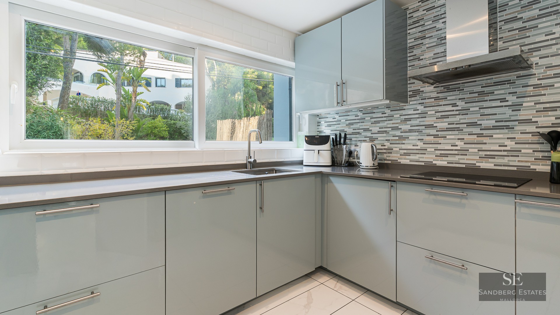 Cuisine moderne avec meubles bleu clair, plans de travail gris, grande fenêtre et crédence en mosaïque.