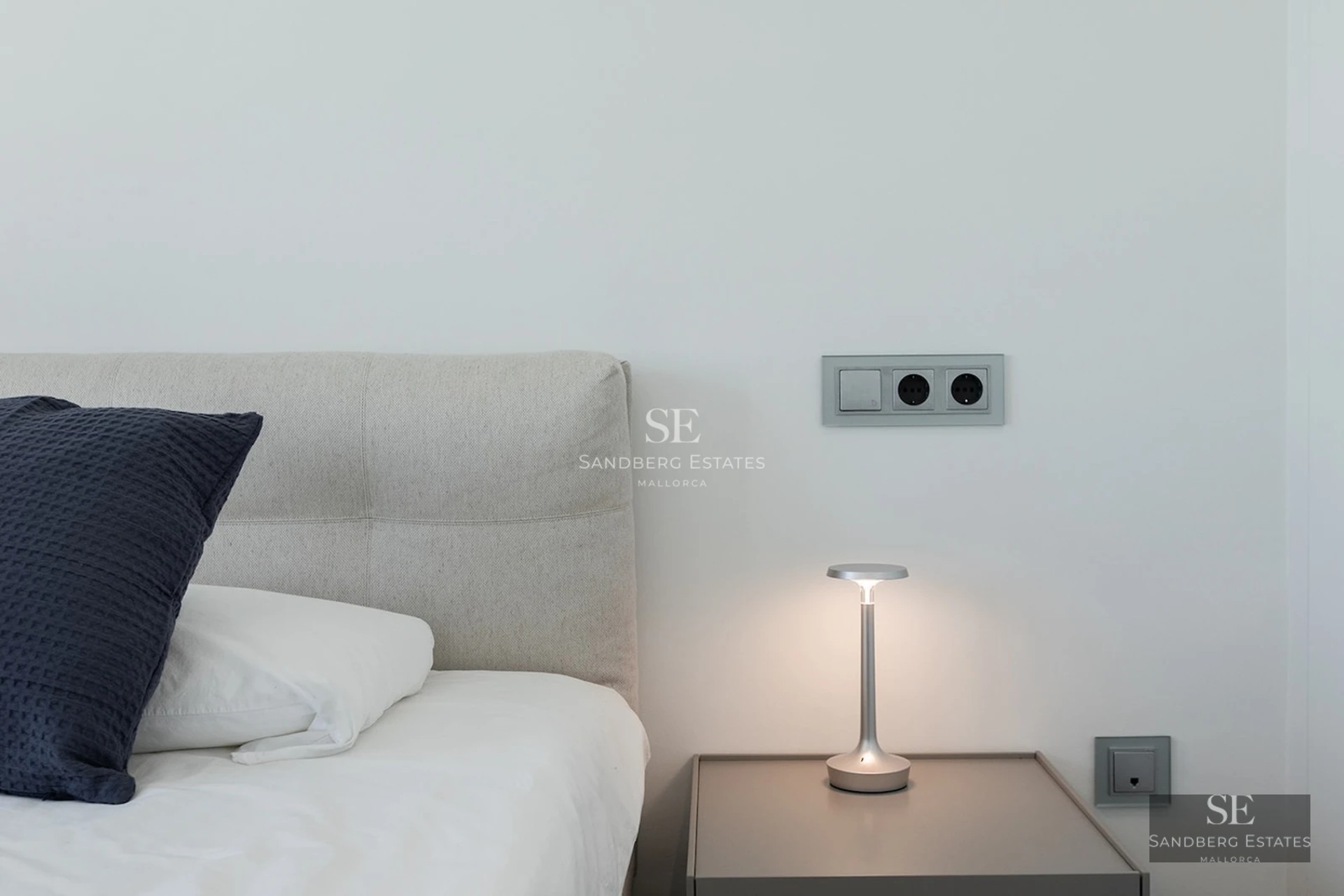 Close-up of a grey headboard, navy pillow, and a sleek silver lamp on a minimalist bedside table.