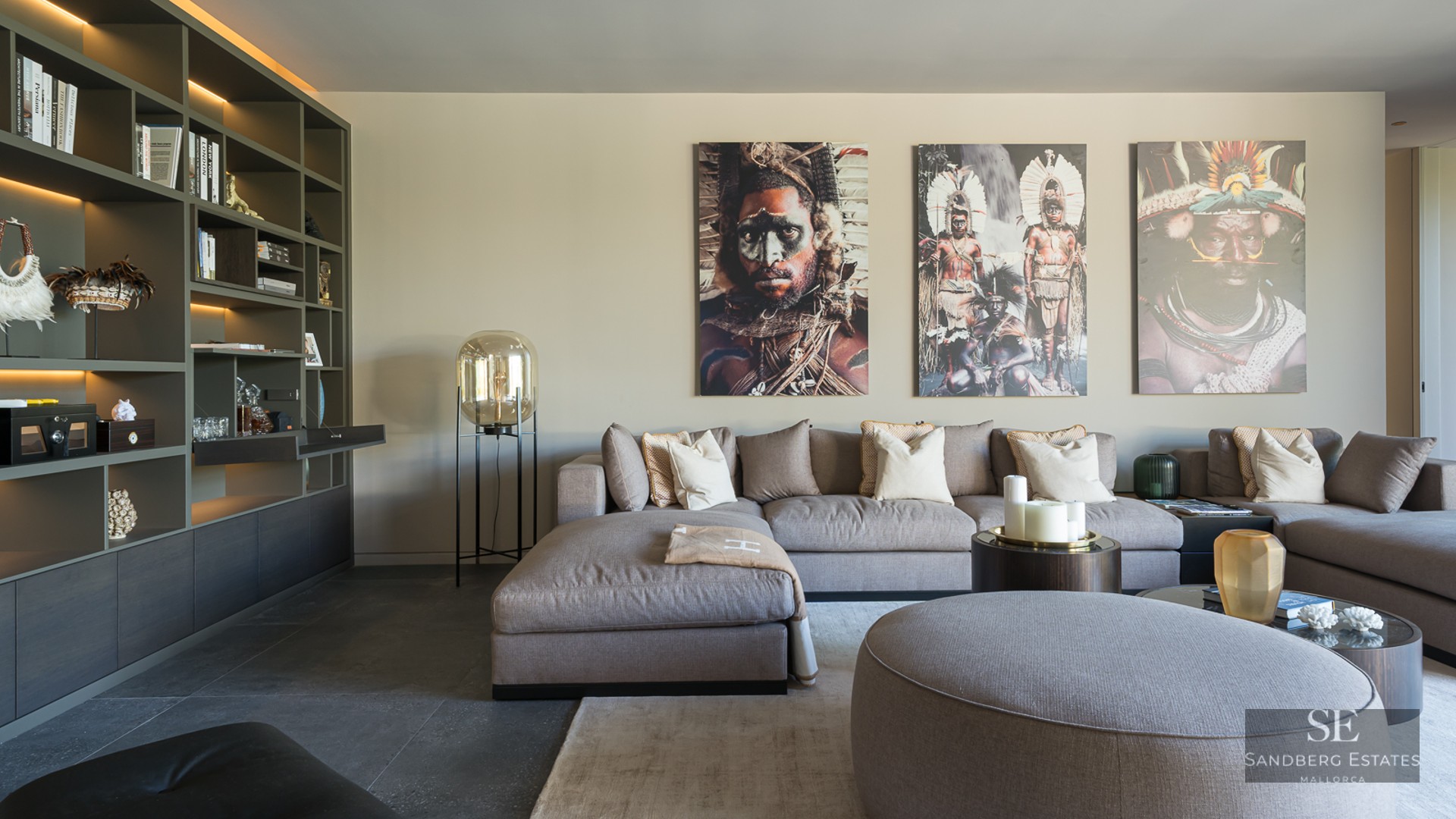 Modern living room with a large grey modular sofa, built-in library, and three large tribal portraits on the wall.