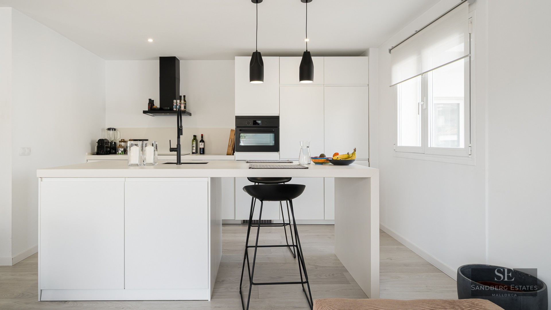 Bright white kitchen featuring a large central island with black bar stools and black hanging pendant lights.