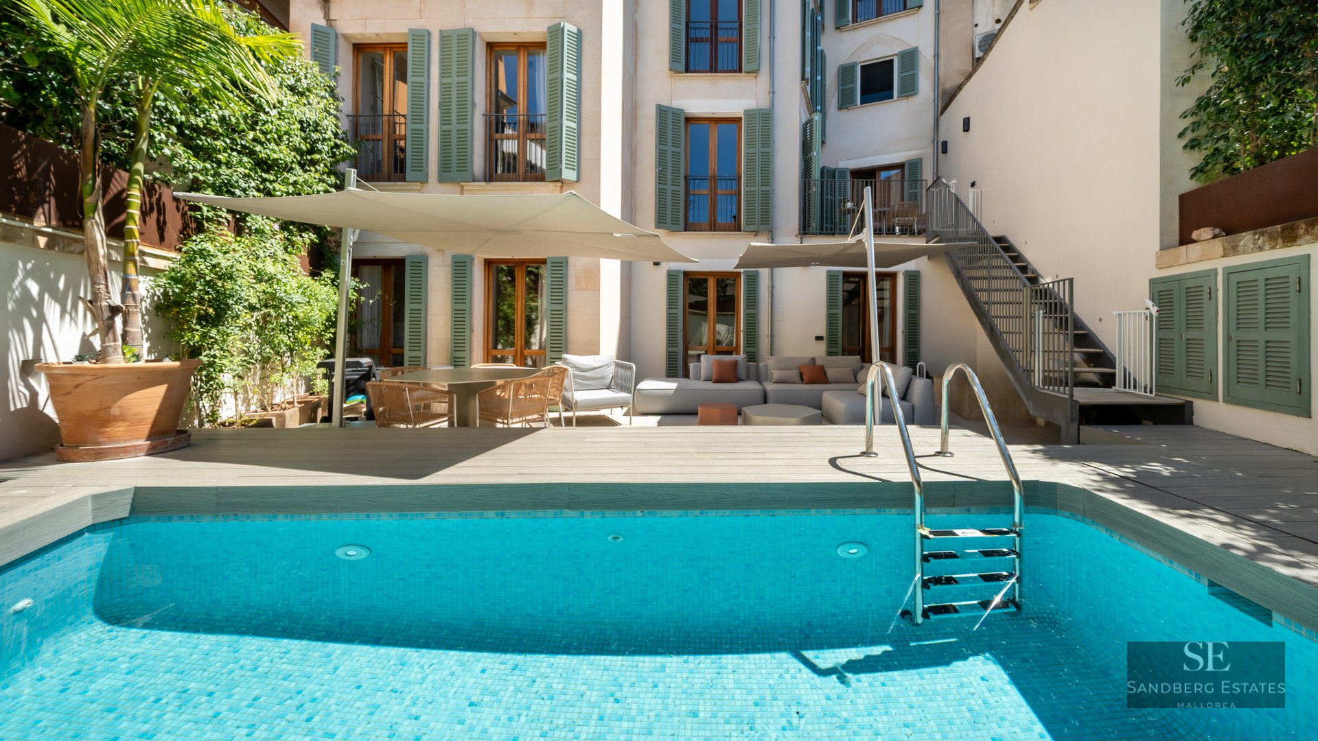 A turquoise swimming pool with a wooden deck, lounge furniture under shade sails, and a building with green shutters.