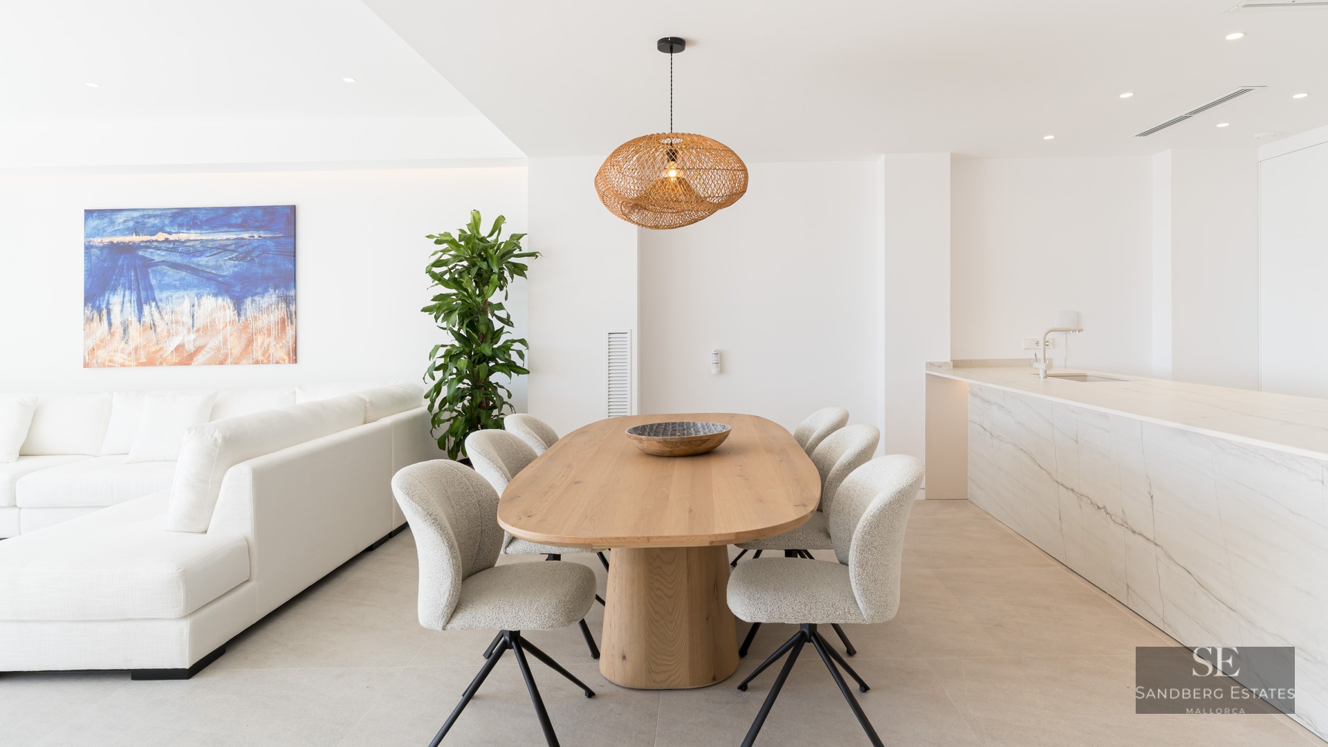 Bright modern dining room with oval wooden table, woven pendant light, and marble kitchen island in an open-plan space.