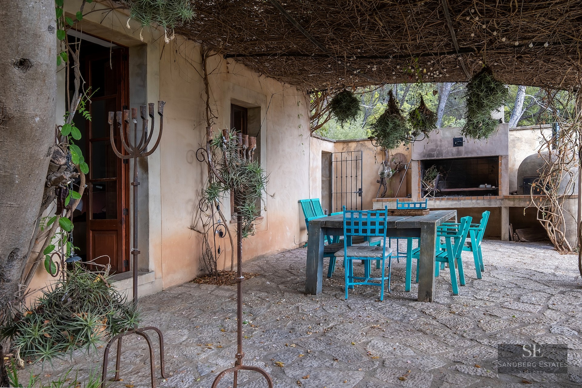 Stone terrace with wooden table, turquoise chairs, thatched roof, and an outdoor barbecue in the background.