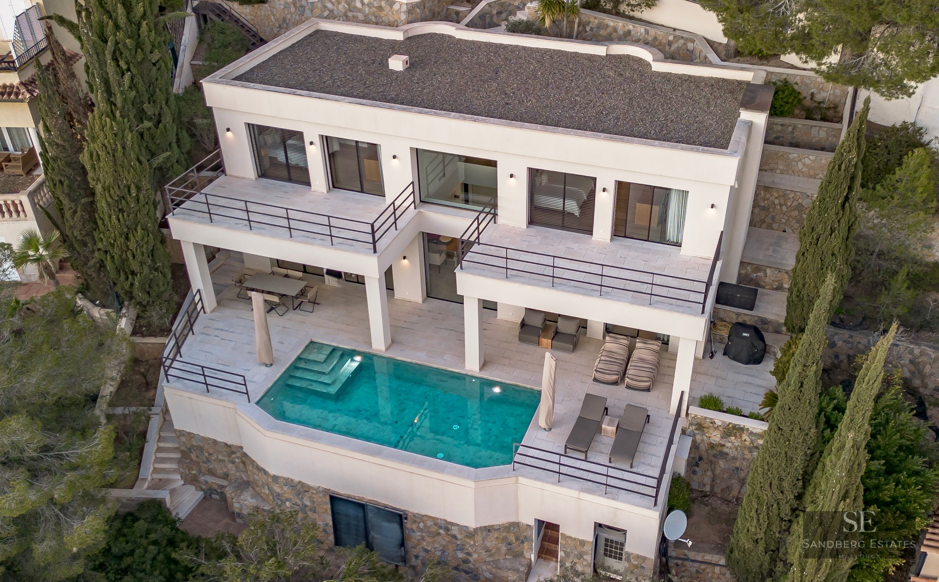 Aerial view of a modern white villa featuring a turquoise pool, multi-level terraces, and cypress trees.