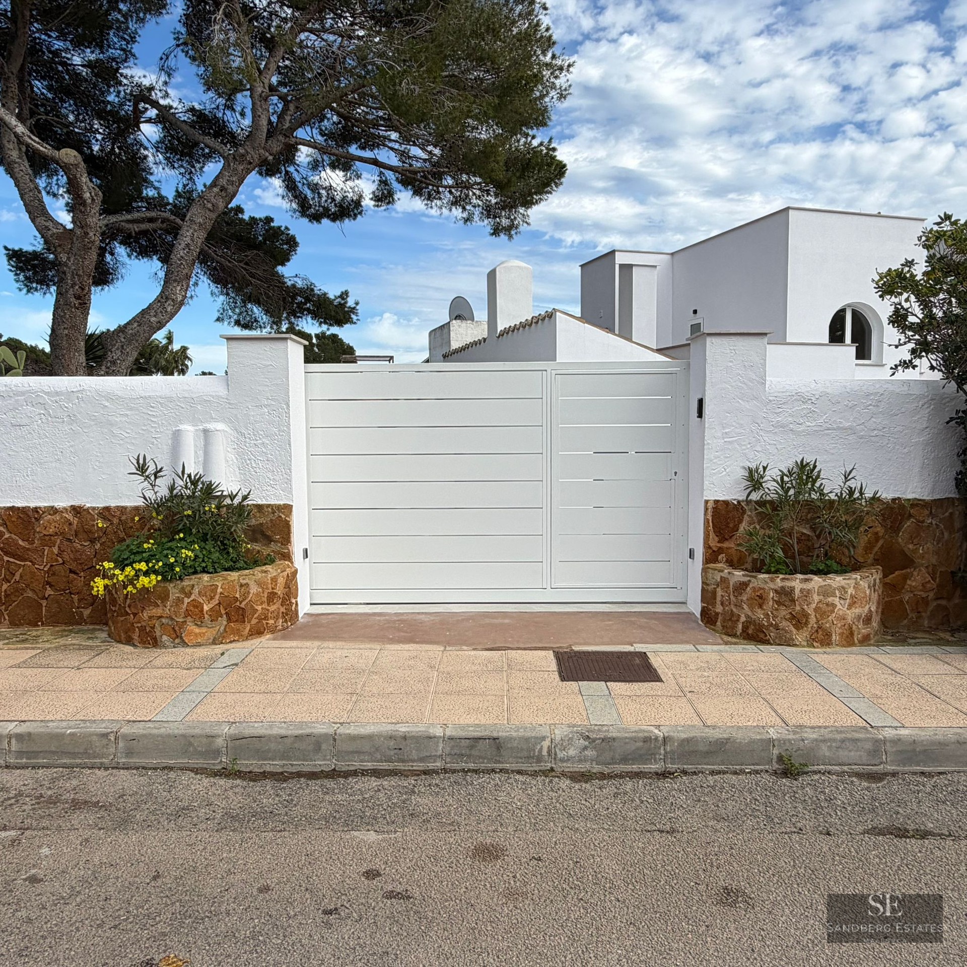 Large white metal gate flanked by white stucco and brown stone walls under a blue sky with a pine tree.