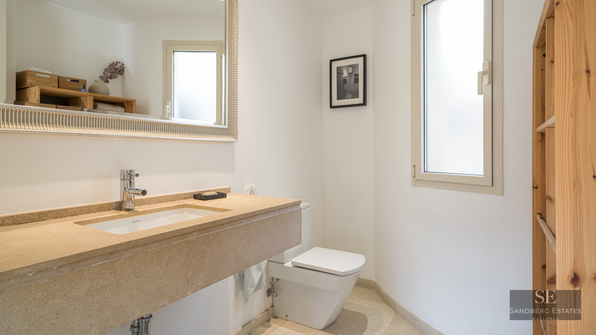 Bright bathroom featuring a tan stone vanity, large silver-framed mirror, white toilet, and frosted window.
