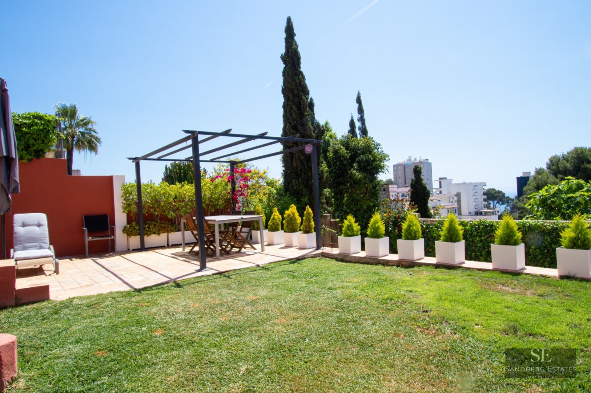 Sunny terrace with a dining table under a metal pergola, a lawn area, and white planters overlooking a city view.