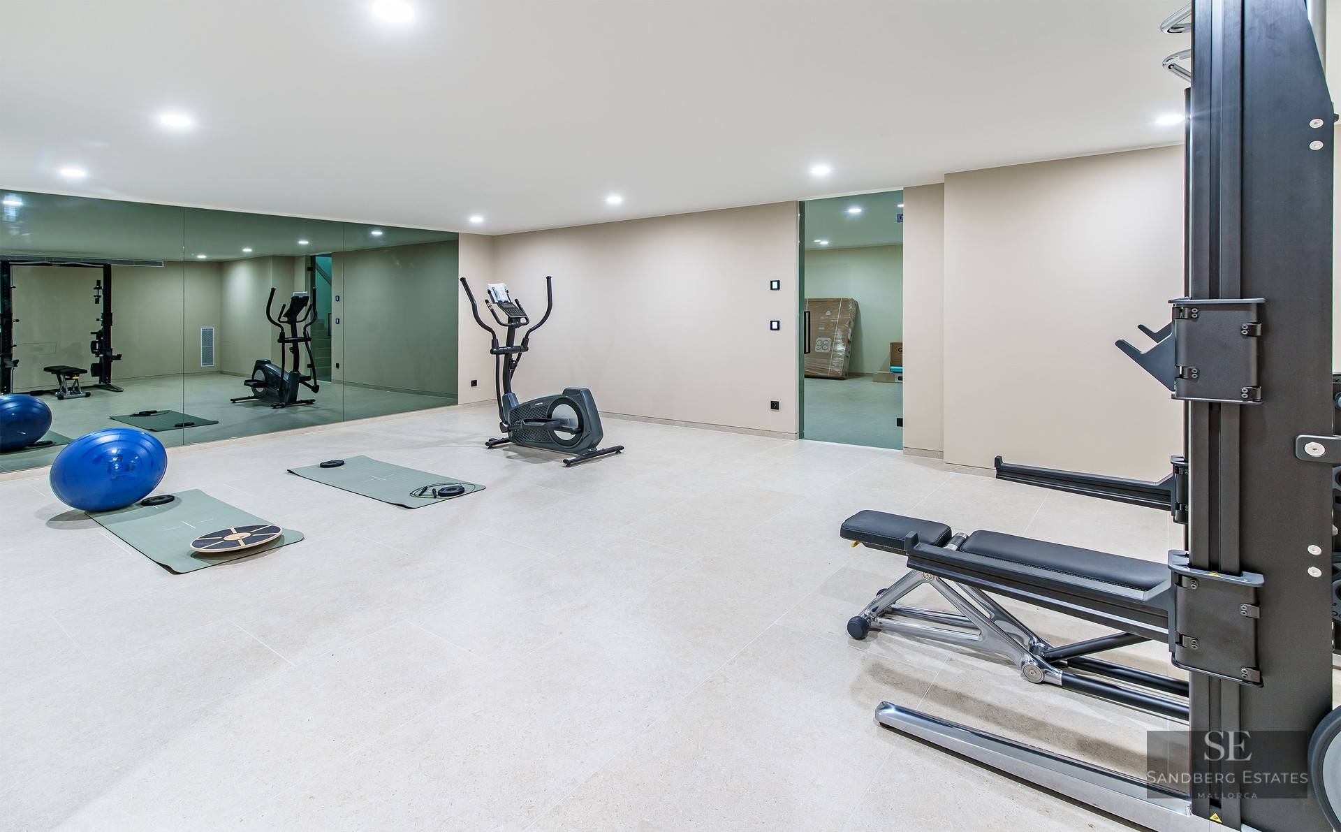 Spacious gym with elliptical trainer, weight bench, mats, and large wall mirrors under bright ceiling lights.