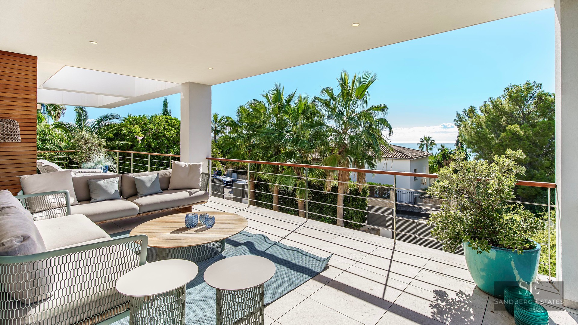 Terrasse moderne avec canapés gris, table en bois et touches turquoise offrant une vue sur les palmiers et la mer.