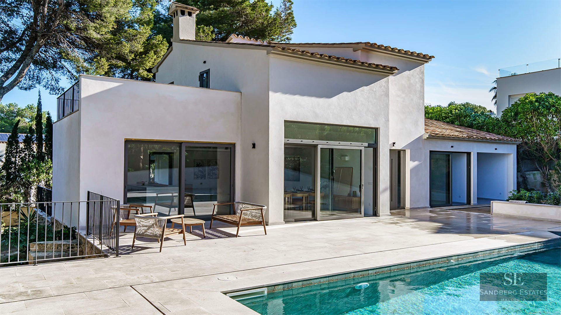 A large turquoise swimming pool in front of a modern white villa with stone terrace and lounge furniture.