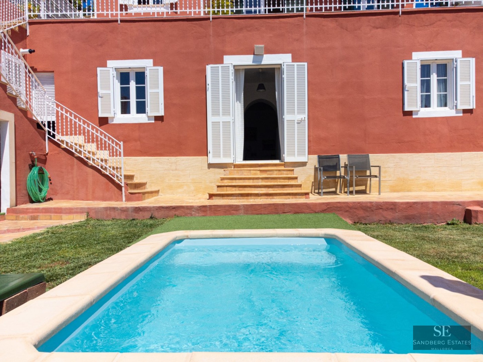 Rectangular turquoise pool in front of a red Mediterranean villa with white shutters and stone terrace.
