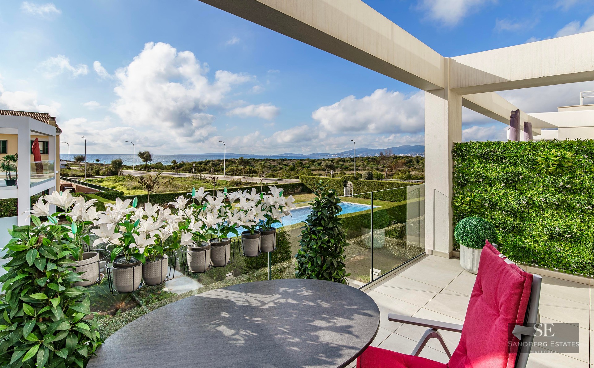 Luxury terrace with a round table, red chair, and glass railing overlooking a garden, pool, and the sea.