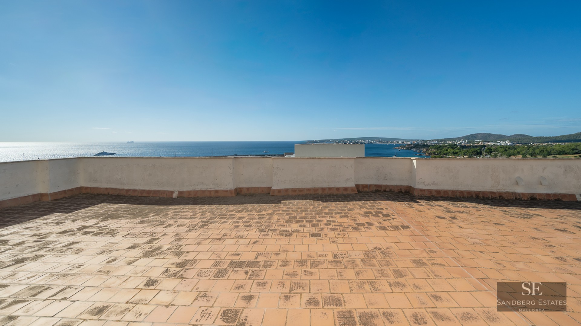 Large rooftop terrace with terracotta tiles overlooking a clear blue sea and coastline under a bright sky.