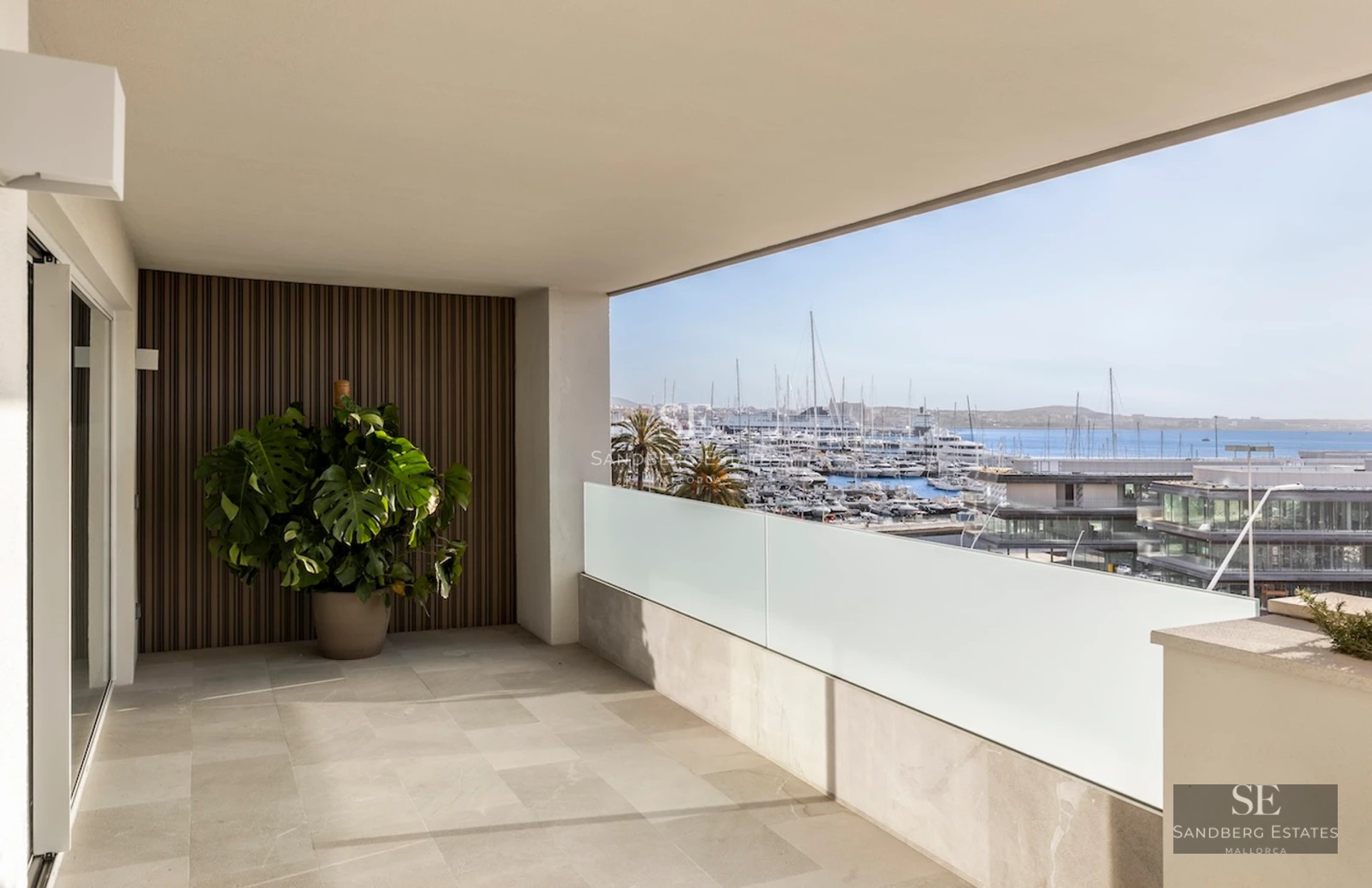 Spacious balcony with sea view. Glass railing, wooden floor, modern furniture, and lush vegetation.