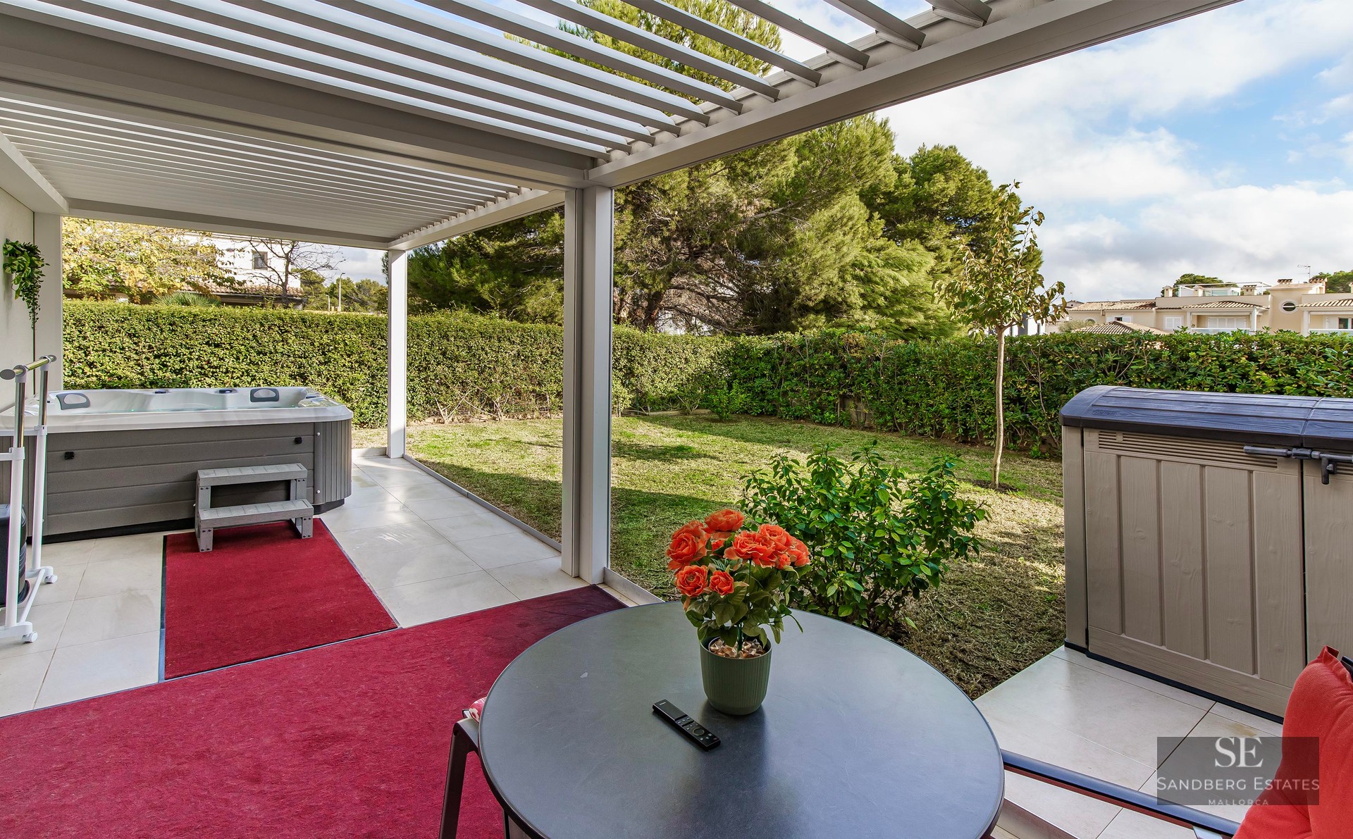 Outdoor terrace with a white pergola, grey hot tub, red rugs, and a view of a green garden with hedges.