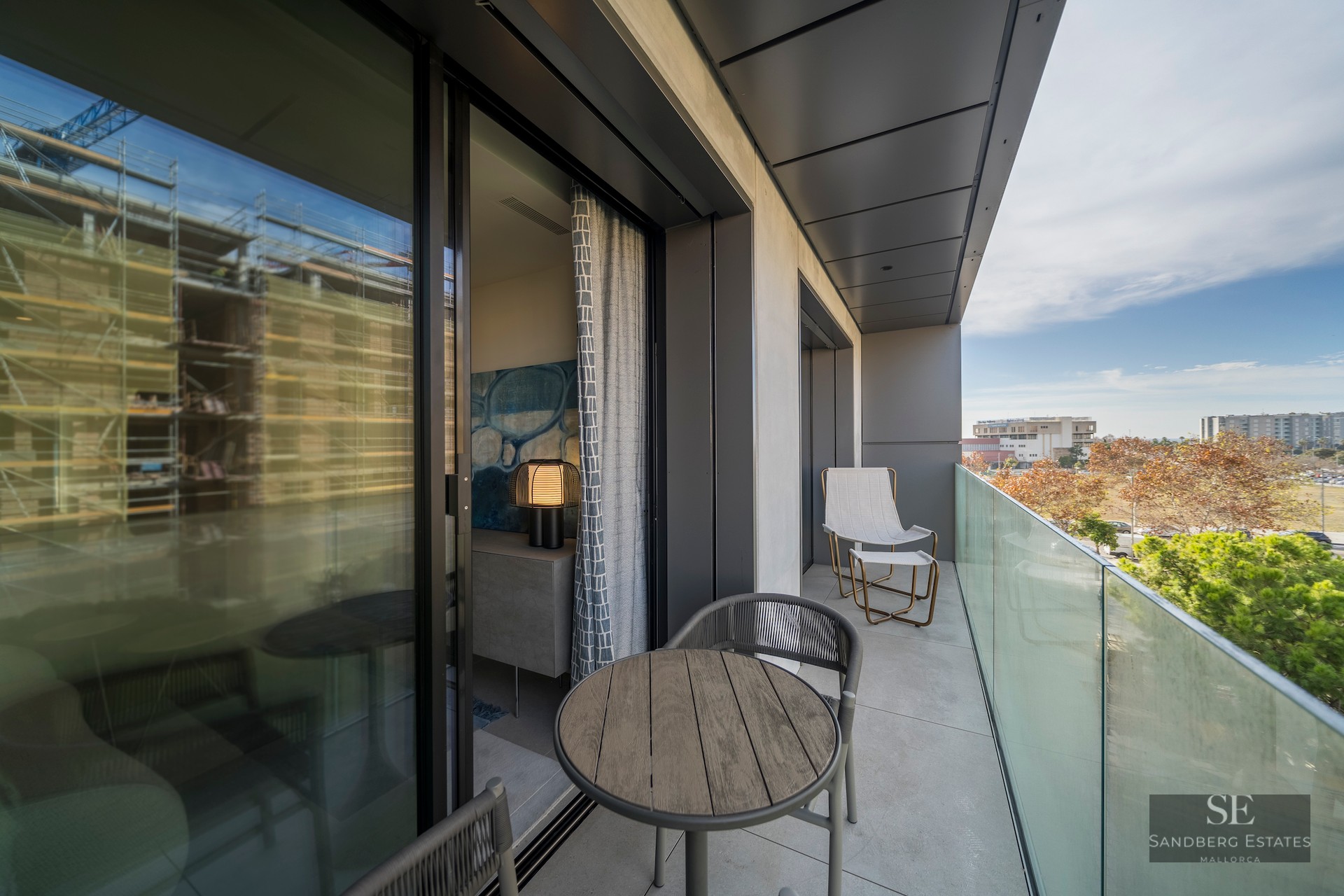 A contemporary balcony with a glass railing, wooden table, and comfortable seating overlooking a green park area.