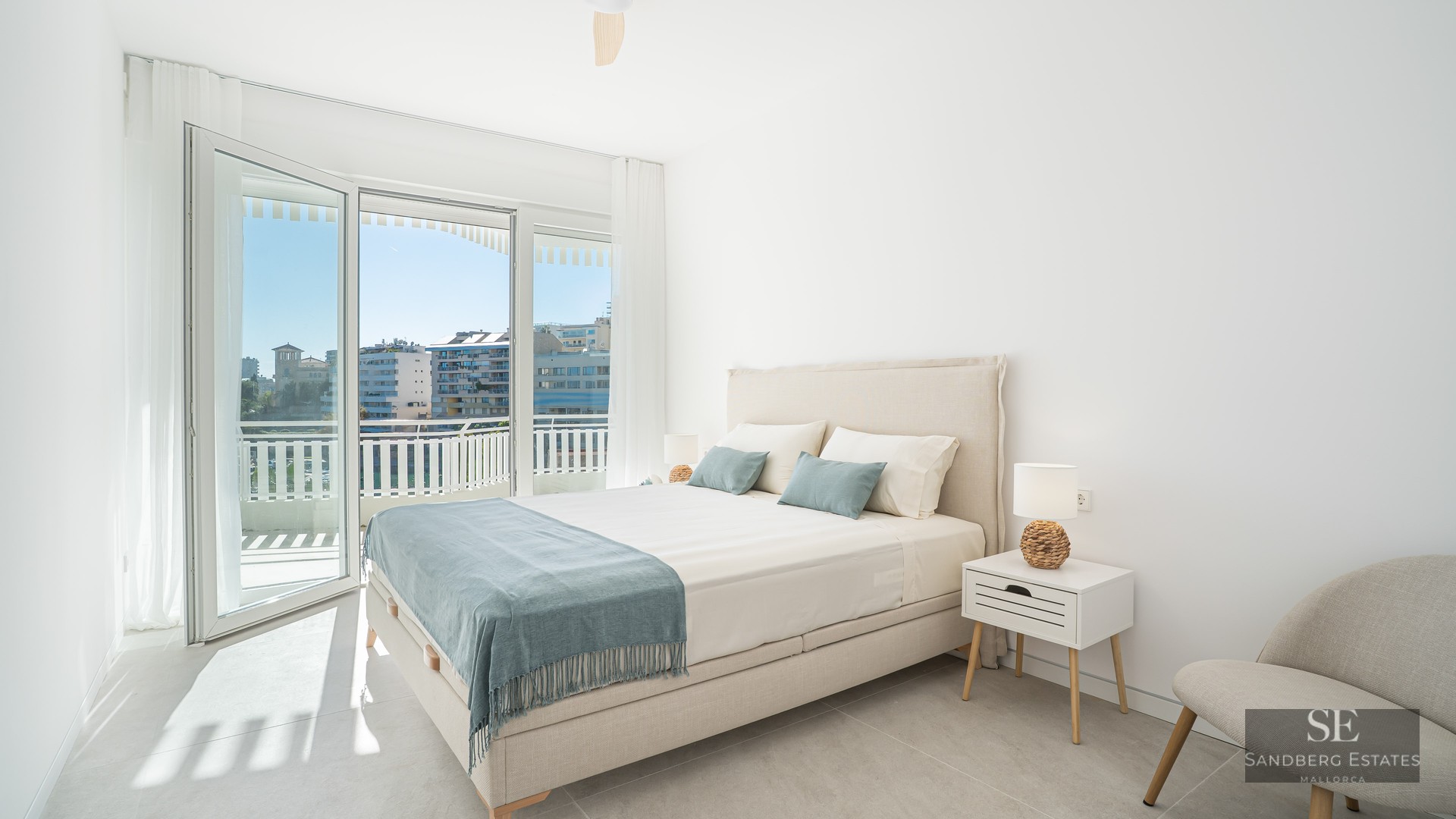 Bright bedroom with a double bed, bedside tables, and large glass doors opening to a balcony with a city view.